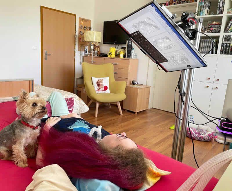Petra leží na posteli, sleduje monitor, pri nej pes. Vedľa postele je umiestnený stojan držiaci monitor, ktorý sa nachádza zhruba pol metra nad Petrou.