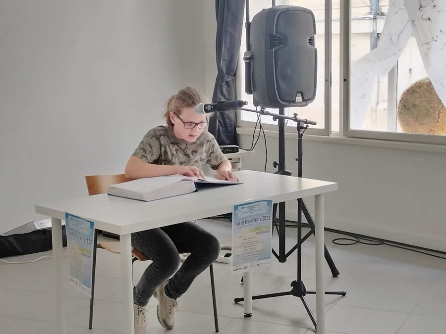 Za stolom s mikrofónom sedí Viktor Radošovský, číta z brailovej knihy. Vedľa stola sa nachádza stojan s reproduktorom.