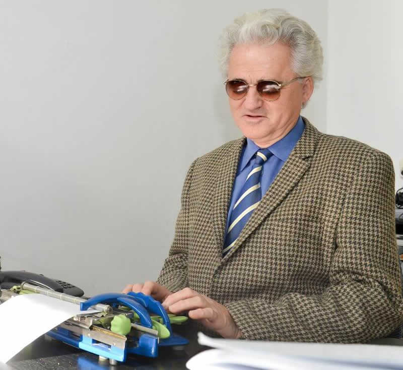 Branislav Mamojka v obleku sedí za stolom a pracuje na Pichtovom stroji pre písanie Braillovho písma.
