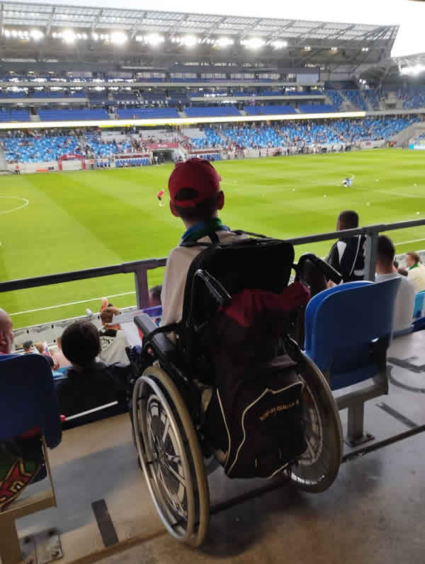 Tadeáš sediaci vo vozíku v hľadisku sleduje dianie na futbalovom ihrisku, vedľa neho je miesto vyhradené pre sprevádzajúcu osobu.