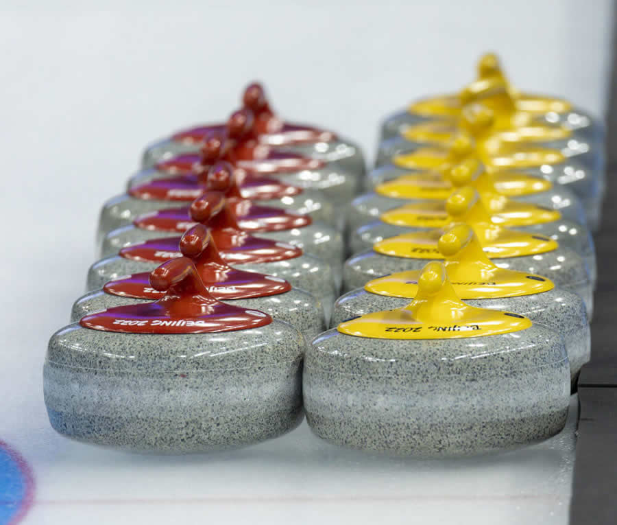 Detailný záber na dva rady curlingových kameňov uložených na ľade, rad kameňov vľavo má červené rúčky, rad vpravo žlté.