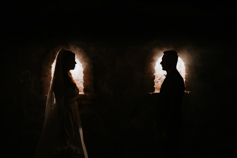 Muž a žena vo svadobných šatách s kyticou v ruke stoja oproti sebe v polotmavej starej miestnosti s dvomi osvetlenými oknami, ktoré tvoria pozadie siluet ich postáv.